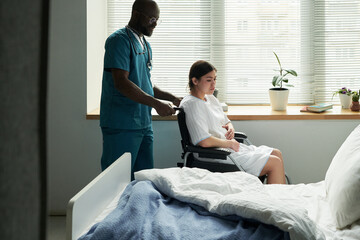 Black male nurse assisting young pregnant Caucasian woman with disability in wheelchair,...