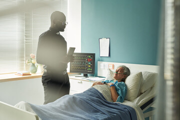 Senior Caucasian woman lying in hospital bed receiving care while Black male priest with Bible...