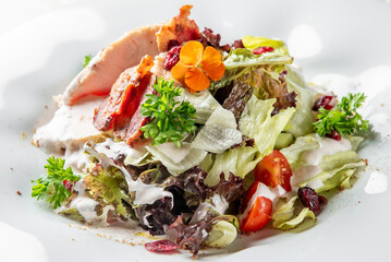 Fresh chicken salad with mixed greens, cherry tomatoes, cranberries, bacon, creamy dressing, and edible flower garnish on a white plate.