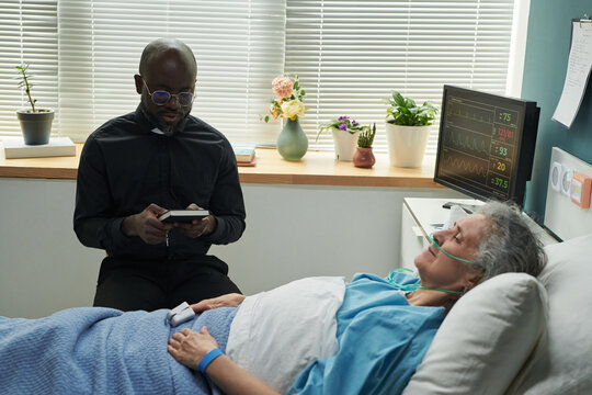 Middle aged Black male chaplain sitting beside hospital bed reading Bible while senior Caucasian woman lying under blanket with oxygen tube and monitoring device, during palliative care in hospice