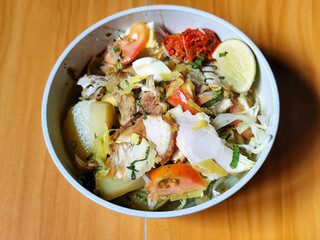 A bowl of Indonesian chicken soto with shredded chicken, sliced boiled egg, vermicelli, sliced cabbage, bean sprouts, celery, chili sauce and lime, mixed with delicious sauce. On a wooden table.	

