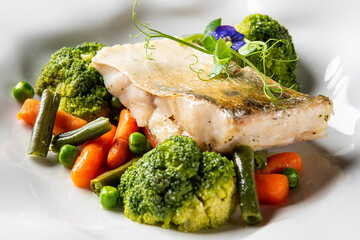 Grilled fish fillet with microgreens and edible flower, served with steamed broccoli, baby carrots, green beans, and peas on a white plate