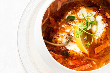 Close-up of traditional soup with vegetables, lemon wedge, sour cream, and herbs in a white bowl on a patterned plate, rich reddish broth, top view