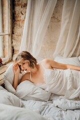 a blonde woman in a white dress on white sheets by the window