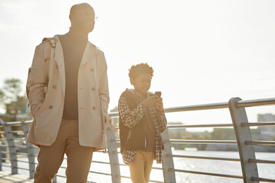 Horizontal medium long low angle shot of Black father and son strolling along river embankment enjoying spare time together, boy surfing Internet on smartphone