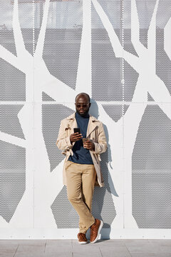 Vertical shot of handsome young adult Black man wearing stylish outfit standing relaxed against building with decorative panel facing surfing Internet on smartphone