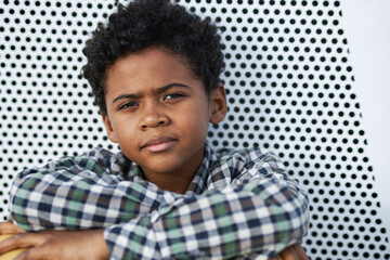 Horizontal close-up portrait of handsome Black boy wearing checked shirt sitting outdoors against wall looking at camera