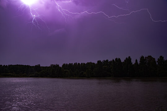 Bright purple lightning streaks across a stormy night sky above a dark forest and calm lake. - Powered by Adobe