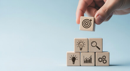 Wooden cubes with business icons, hand placing a target cube on top, light blue background, showcasing goal achievement and strategic planning