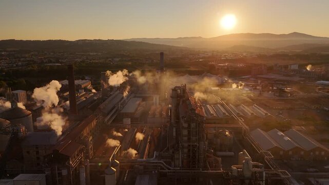 Drone view of vast industrial factory with towering smokestacks, expansive grounds, and glowing sunset sky, highlighting industrial scale and atmospheric evening mood