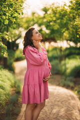 Beautiful and happy pregnant young woman in pink dress on the path in the park