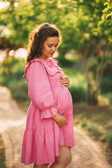 Beautiful and happy pregnant young woman in pink dress on the path in the park
