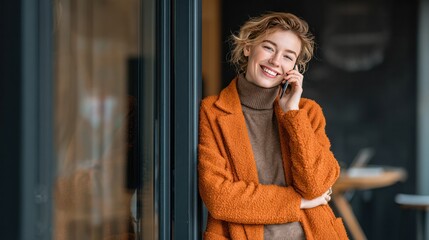 Fototapeta premium Stylish Woman on Phone: A confident woman with short, wavy blonde hair smiles warmly while talking on her mobile phone, leaning against a modern doorway.