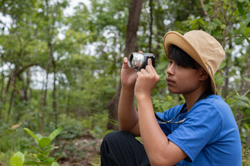 Asian teen boy in blue t-shirt and wears hat and holding camera for exploring the forest on a sunny day, enjoying outdoor adventure and wildlife observation.