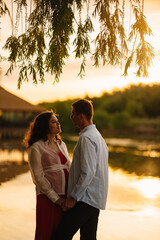 man and pregnant woman in red dress at sunset on the riverbank