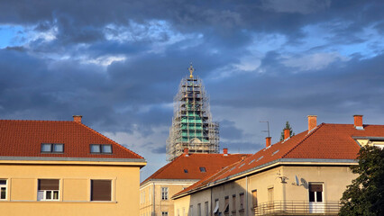 Tall church tower covered in scaffolding dominates apartment rooftops with red tiles, all bathed in...