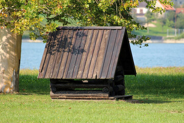 Wooden A-frame log structure sits on grassy shore by blue lake, partially shaded by plane tree,...