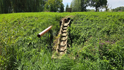 Rural embankment features concrete drainage stairs and three open pipes, surrounded by lush summer...