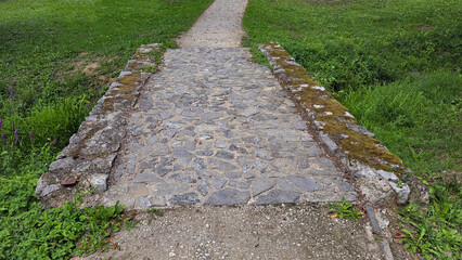 Stone bridge made of irregular rocks and moss crosses shallow ditch, integrating gravel path and...
