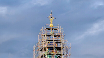 Detailed golden cross and sphere atop church dome, encased in metal scaffolding, rise against...