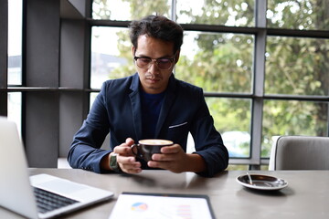 A cheerful young man in casual attire sits indoors, enjoying a hot drink while reading, relaxed in...