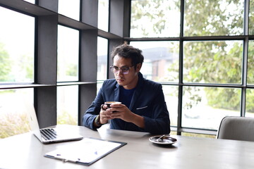 A cheerful young man in casual attire sits indoors, enjoying a hot drink while reading, relaxed in...