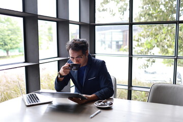 A cheerful young man in casual attire sits indoors, enjoying a hot drink while reading, relaxed in...