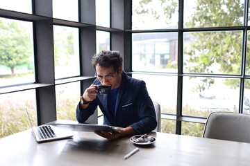 A cheerful young man in casual attire sits indoors, enjoying a hot drink while reading, relaxed in...