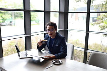 A cheerful young man in casual attire sits indoors, enjoying a hot drink while reading, relaxed in...