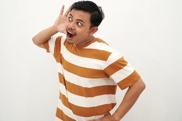Young Asian man in striped tshirt posing with a curious expression, holding one hand behind his ear as if trying to listen closely, standing against a white background