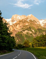 Naklejka premium Winding road curves through lush green valley, majestic snow-capped mountains rise in the background under a partly sunny sky