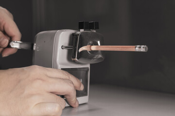 One hand is sharpening a wooden pencil using a grey pencil sharpener.