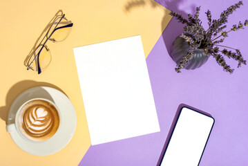 Desktop with blank paper, glasses, coffee cup, mobile phone and lavender flowers in vase on beige and purple background in sunlight. Top view, mockup