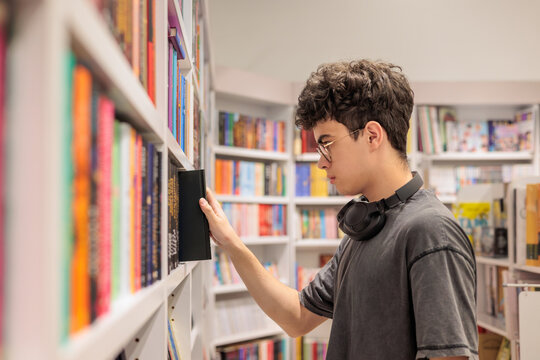 Teenage student with glasses and headphones browsing books in bookstore or library. education, research, academic lifestyle, self development, reading and knowledge
