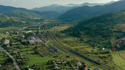 Aerial drone footage of a train moving through scenic mountains near a small settlement. Concept of...