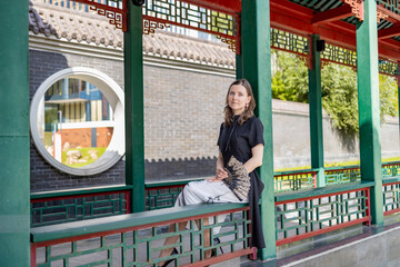 Elegant woman in traditional style black dress with hand fan sitting on a bench in chinese pavilion with green and red columns
