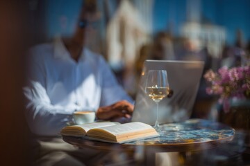 Sunlight bathes a stylish cafe table where a person types on a laptop, with a book and a glass of wine nearby. The relaxing atmosphere invites creativity and contemplation.