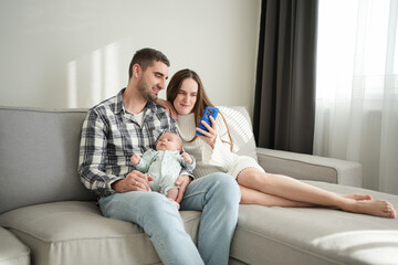 Obraz premium Young family with baby sitting on cozy sofa, smiling and looking at smartphone