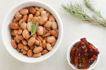 Boiled borlotti beans in a white bowl with a sprig of rosemary and sun-dried tomatoes on a gray background. Italian cuisine. Vegan food. Selective focus. Horizontal orientation. Top view