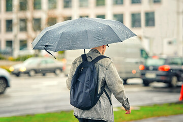 Man walking under black umbrella in heavy rain wearing denim jacket and backpack, facing strong...