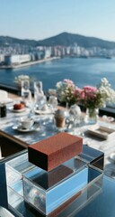 Elevated, dark-brown rectangular object atop a clear acrylic display on a patio table, overlooking cityscape and water