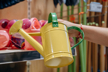 Bright yellow watering can held by a hand, surrounded by colorful gardening tools on a shelf,...