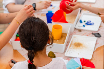 Young girl with dark hair, engaged in creative activities at an after-school program, surrounded by colorful materials and hands of peers, fostering collaboration and artistic expression