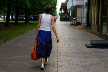 Woman walks down a city sidewalk in a blue skirt and white top during a sunny afternoon in the...