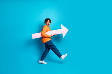 Joyful boy with orange hoodie carries a large arrow while posing against a lively vibrant blue...