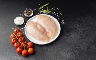 Raw chicken fillets on white plate with salt, tomatoes and herbs on black table