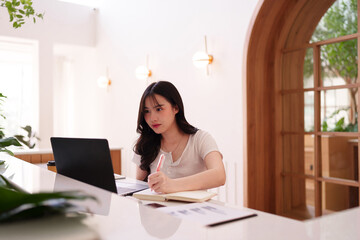 Young Asian woman remote worker using laptop computer focused professional writing notes cafe...