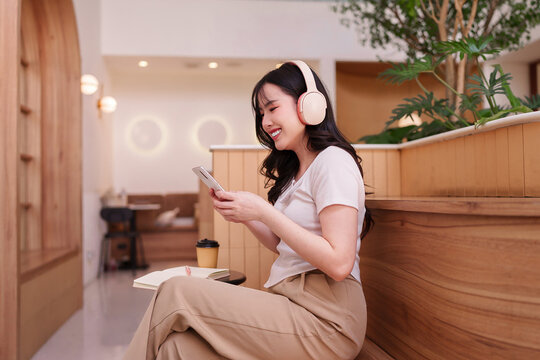 Asian young adult woman happy listening music podcast with headphones using smartphone relaxing modern cafe cozy environment coffee shop digital lifestyle communication technology