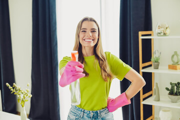Cheerful young woman in casual attire with gloves and a spray bottle performing household cleaning...