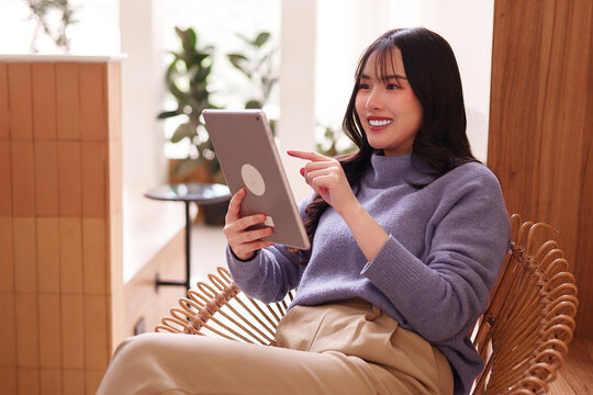 Adult Asian woman happily uses digital tablet modern cafe comfortable rattan chair remote work casual tech connectivity productivity internet browsing
